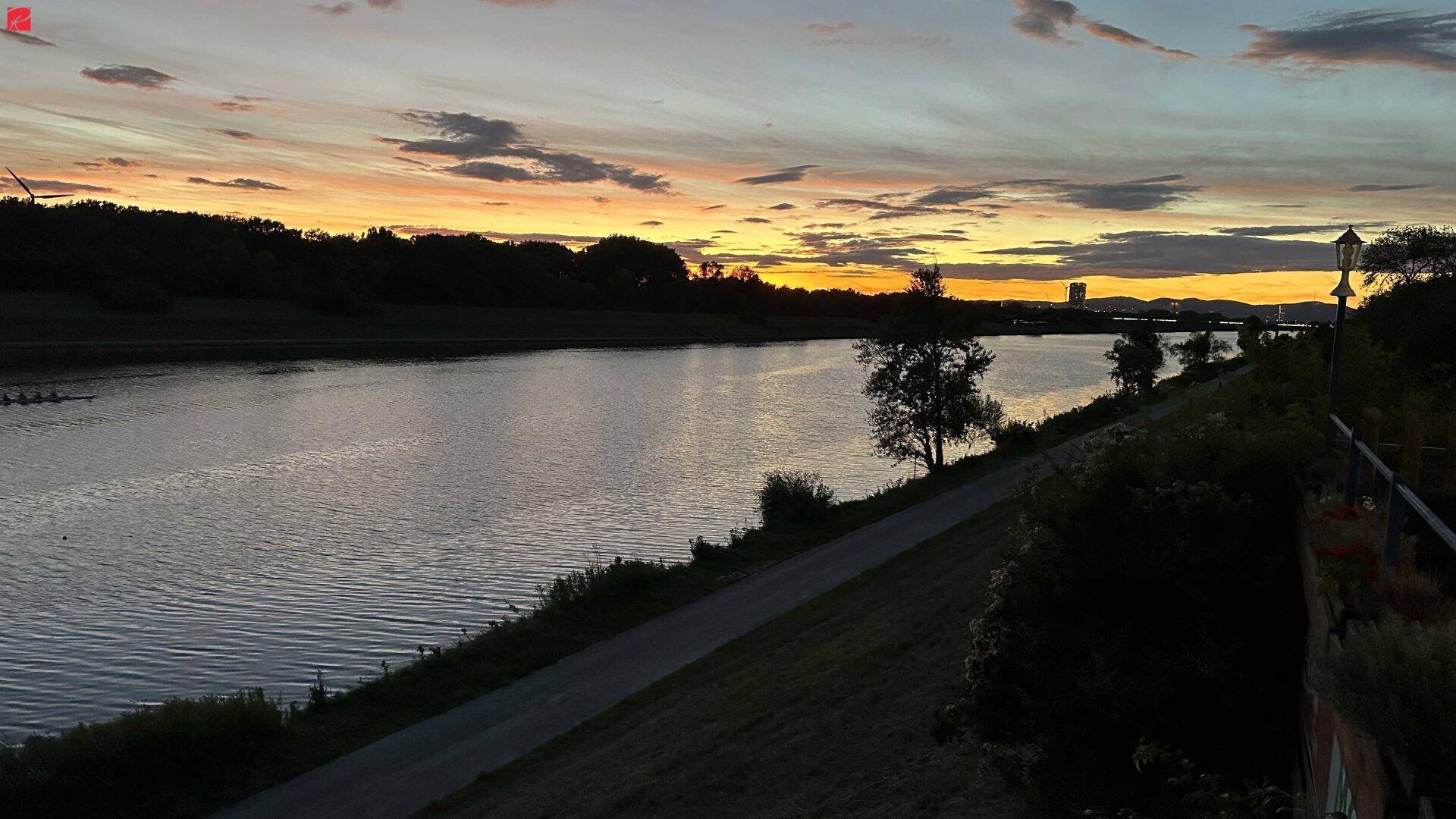 Ruhiger Flusslauf bei Sonnenuntergang mit farbenfrohem Himmel und Uferweg am Horizont.