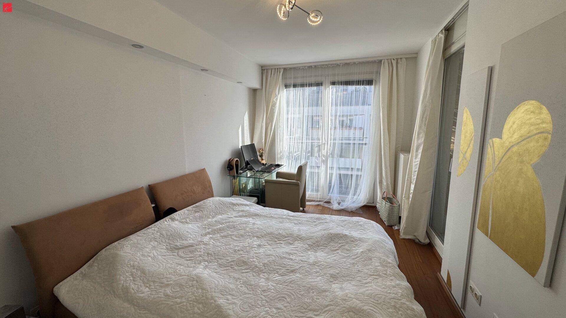 Schlafzimmer mit großem Bett, Schreibtisch und Zugang zu einem Balkon durch bodentiefe Fenster.