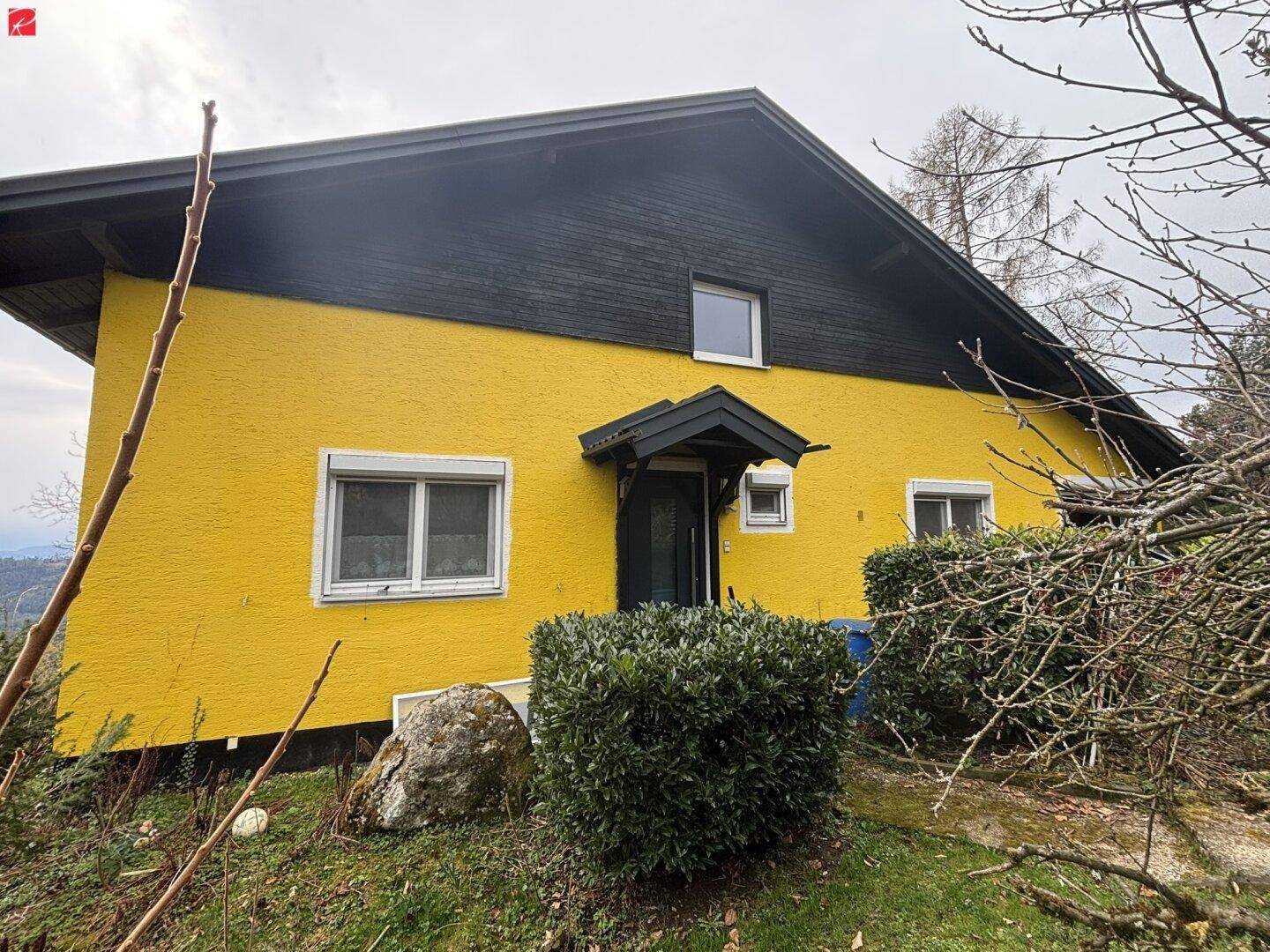 Seitenansicht des gelben Hauses mit schwarzem Dach und gepflegter Bepflanzung im Vorgartenbereich.