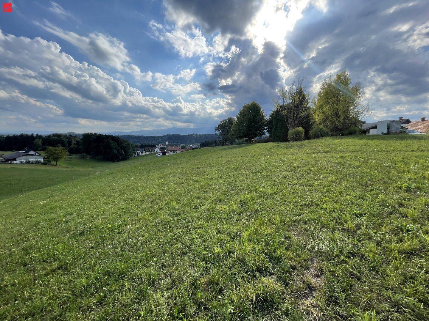 Sonnige Wiesenfläche mit Blick auf eine ländliche Umgebung und dramatischen Wolkenformationen.