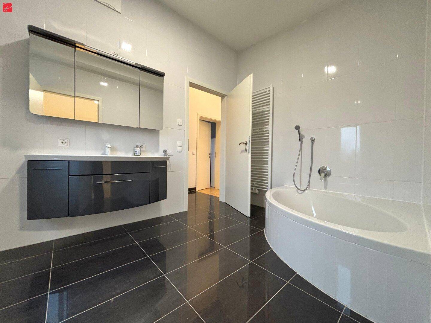 Modernes Badezimmer mit dunklen Bodenfliesen, weißer Wand und Eckbadewanne.