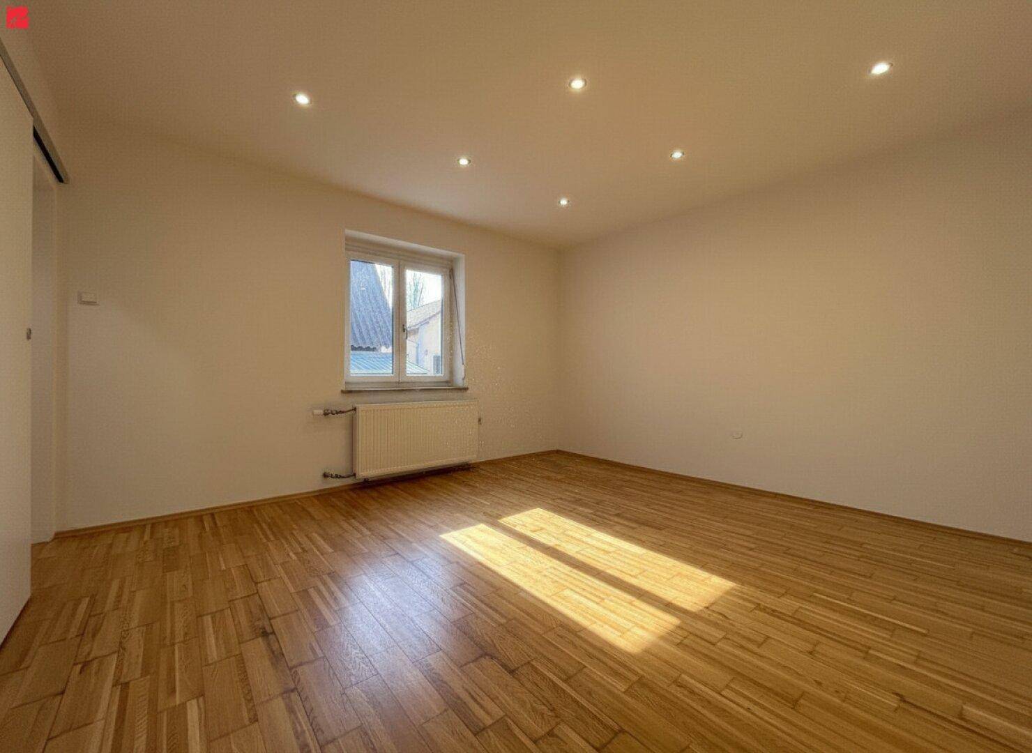 Helles Zimmer mit Holzparkett, Einbaustrahlern und großem Fenster mit viel Sonnenlicht.