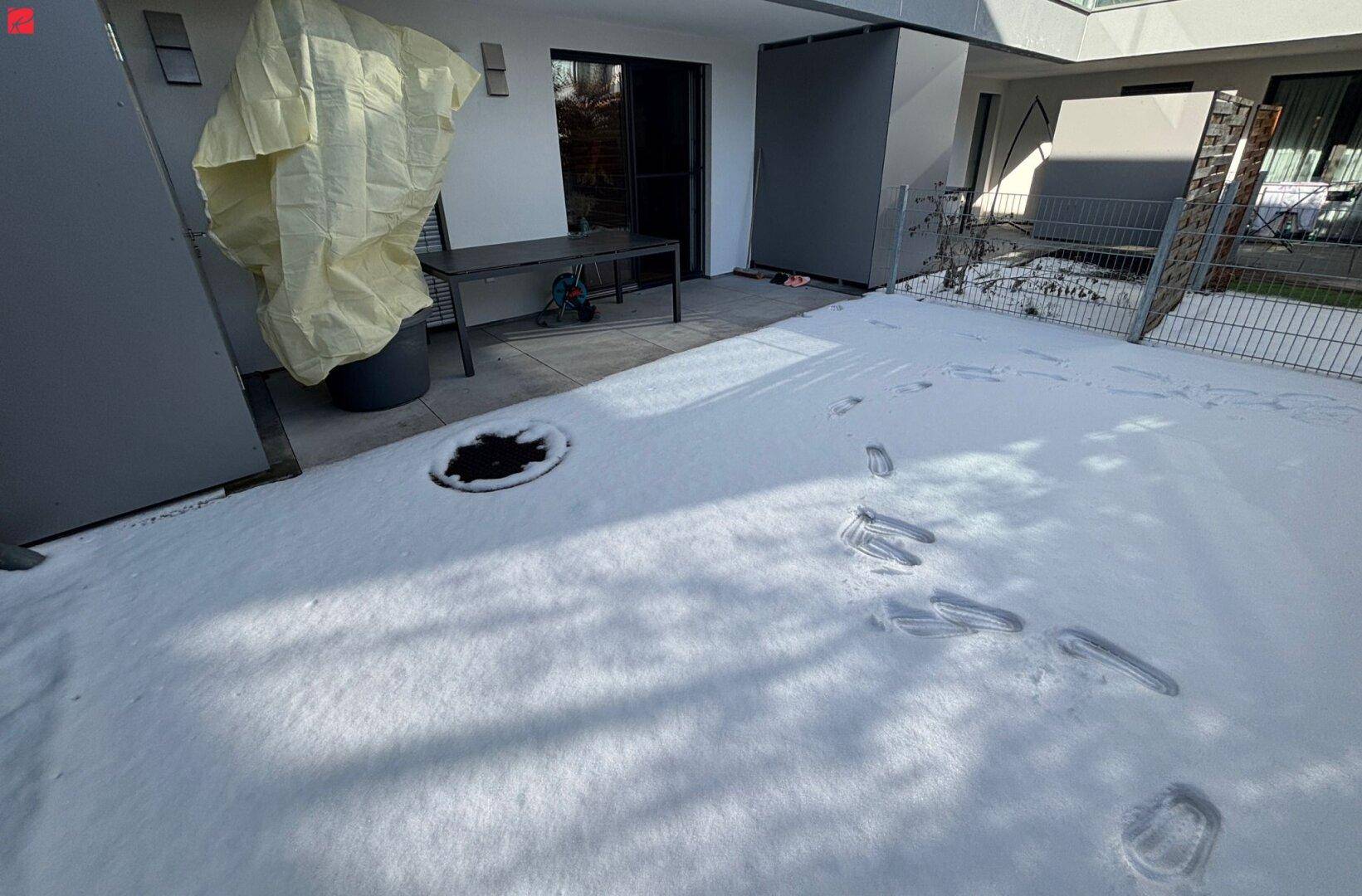 Winterliche Terrasse mit Schnee, Sitzgelegenheiten und Zugang zum Gartenbereich.