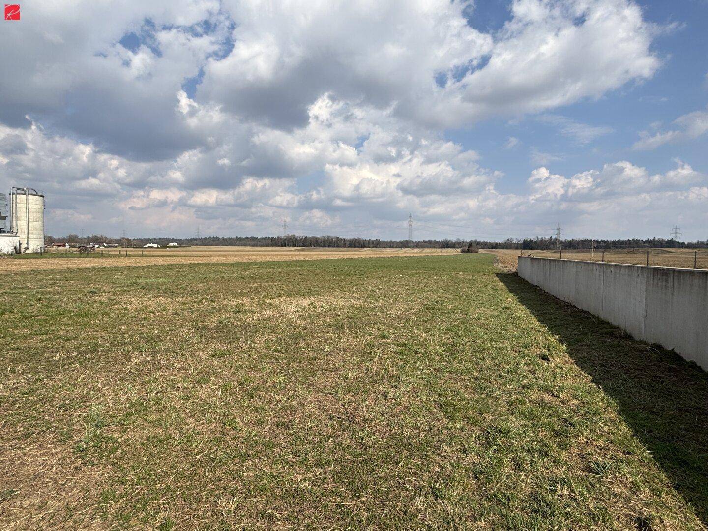 Offene Landschaft mit weiten Feldern und einem Betonzaun im Vordergrund unter einem bewölkten Himmel.