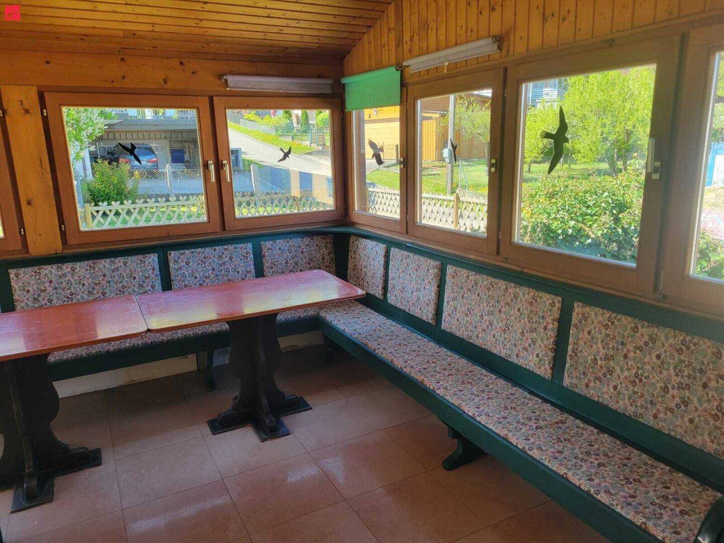 Gemütlicher Wintergarten mit Eckbank, Holztischen und vielen Fenstern für viel Tageslicht.