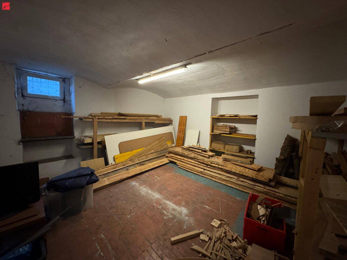Kellerraum mit gelagerten Holzbrettern, Regalen und einem kleinen Fenster, ideal als Abstellraum.