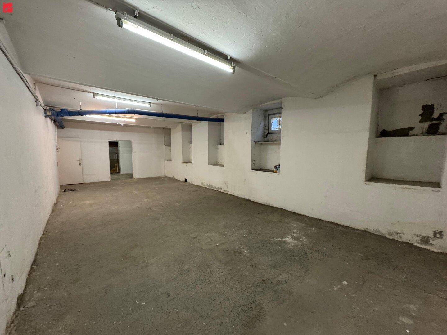 Leerer Kellerraum mit Betonboden, kleinen Fensternischen und sichtbaren blauen Versorgungsrohren an der Decke.