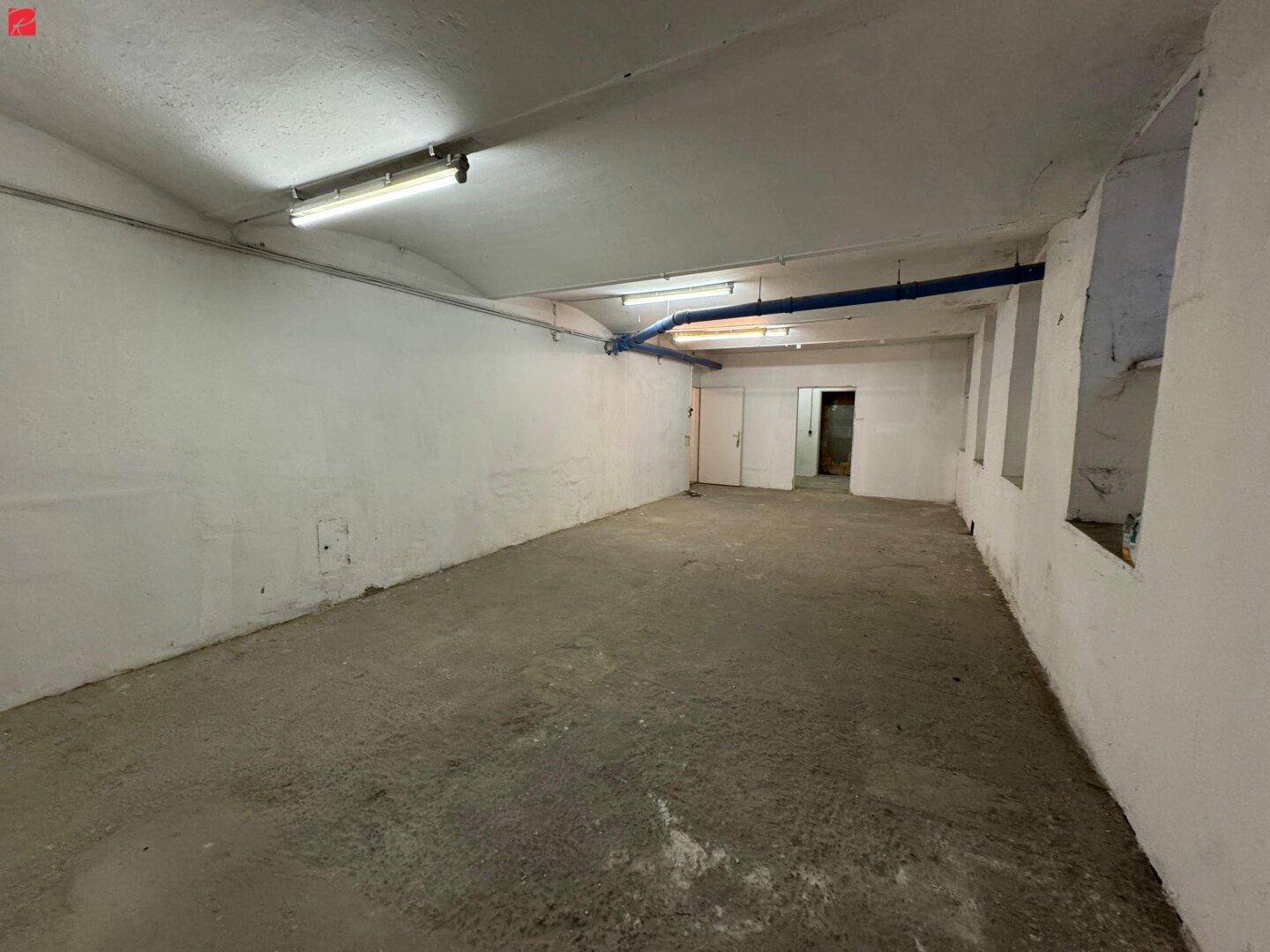 Geräumiger, leerer Kellerraum mit Betonboden, weißen Wänden und sichtbaren blauen Rohren an der Decke.
