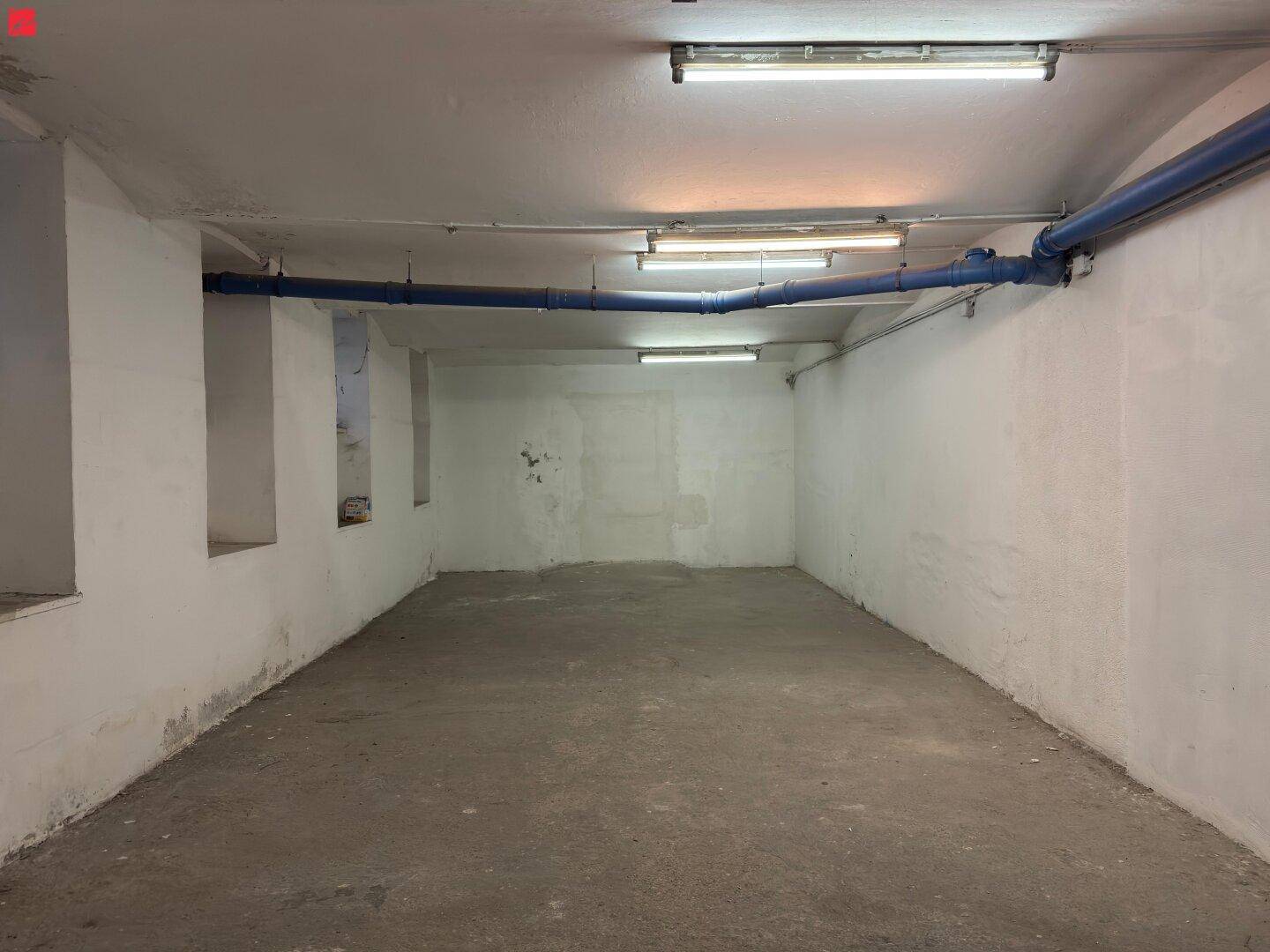 Großer, leerer Kellerraum mit Betonboden, weißen Wänden und markanten blauen Rohren unter der Decke.