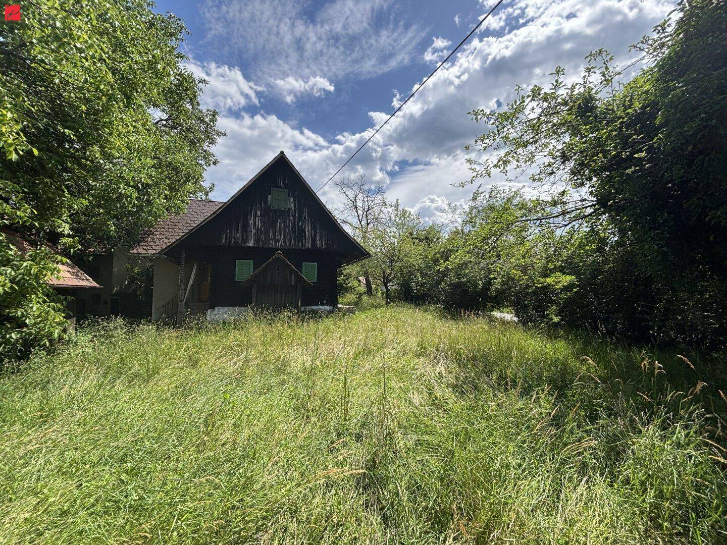 Kleines, dunkles Holzhaus inmitten von hohem Gras und Bäumen unter einem weiten Himmel.