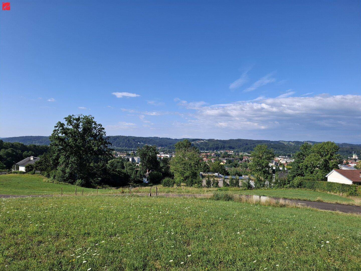 Weitläufige grüne Landschaft mit Blick auf eine Siedlung und bewaldete Hügel unter blauem Himmel.