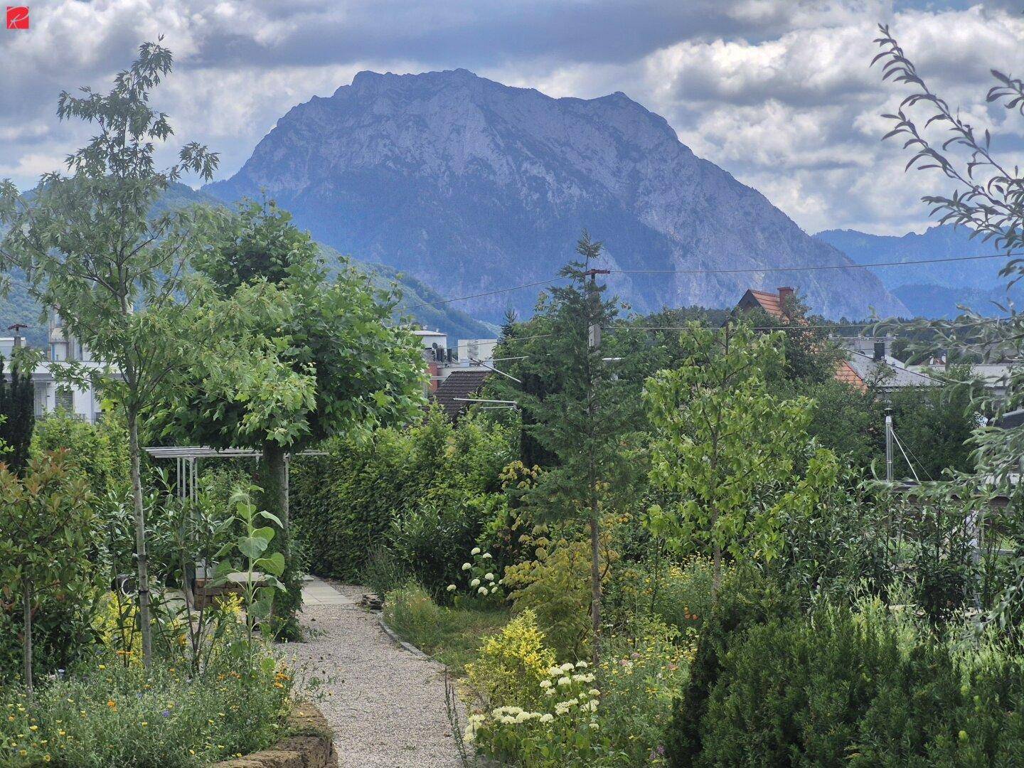 Ein idyllischer Gartenweg führt durch üppiges Grün mit einem majestätischen Bergblick im Hintergrund.