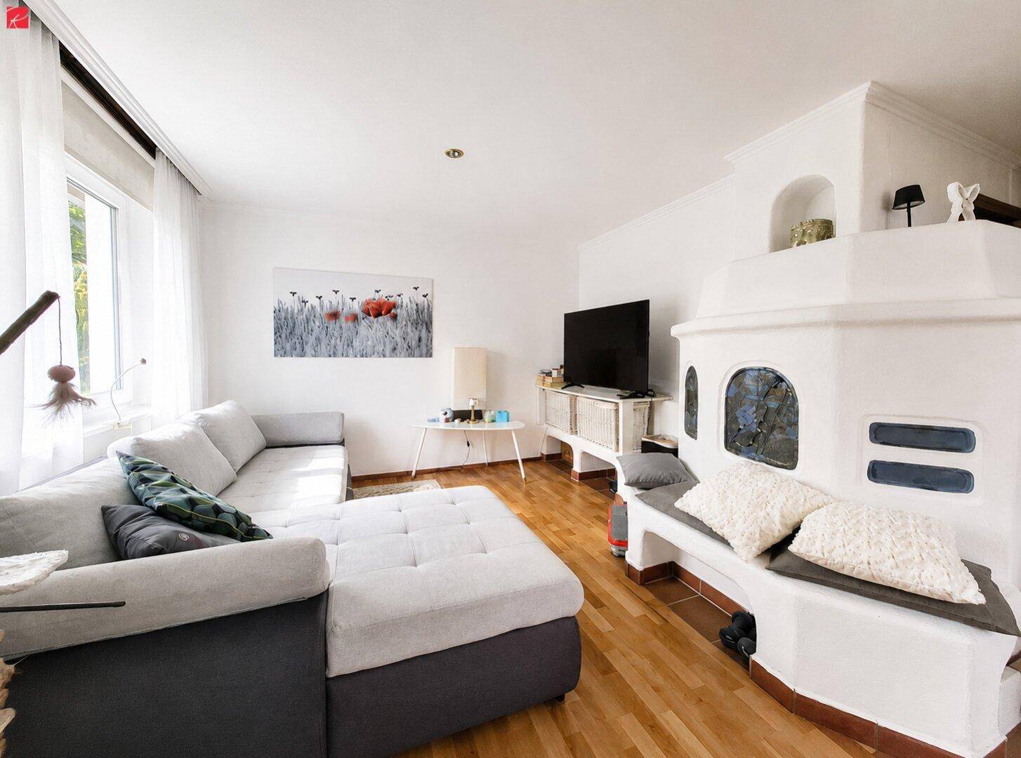 Helles Wohnzimmer mit Ecksofa, Holzboden und traditionellem Kachelofen als gemütlicher Mittelpunkt.