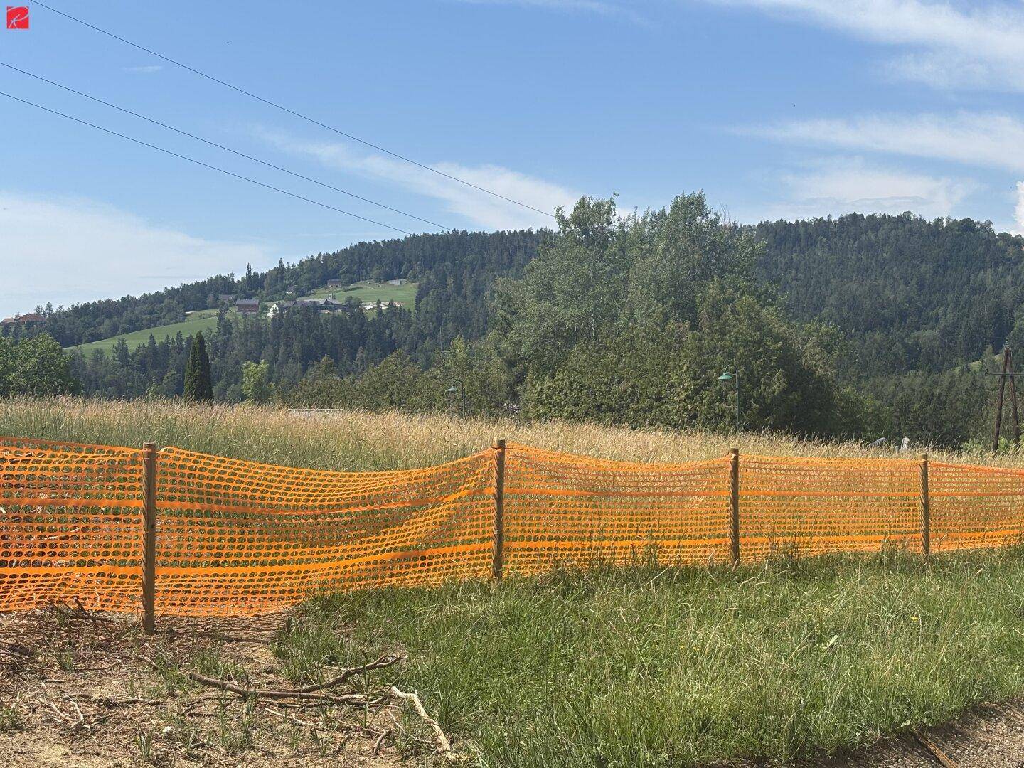 Grüne Landschaft mit Hügeln und Bäumen im Hintergrund, abgegrenzt durch einen orangefarbenen Bauzaun.