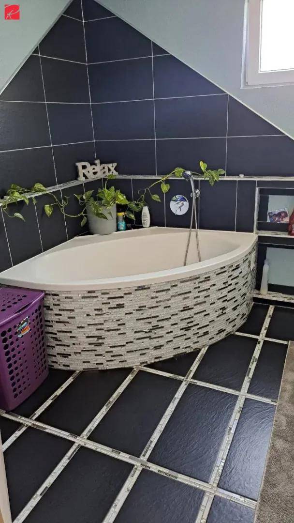 Badezimmer mit einer Eckbadewanne, dunklen Fliesen und einem Fenster, das natürliches Licht hereinlässt.