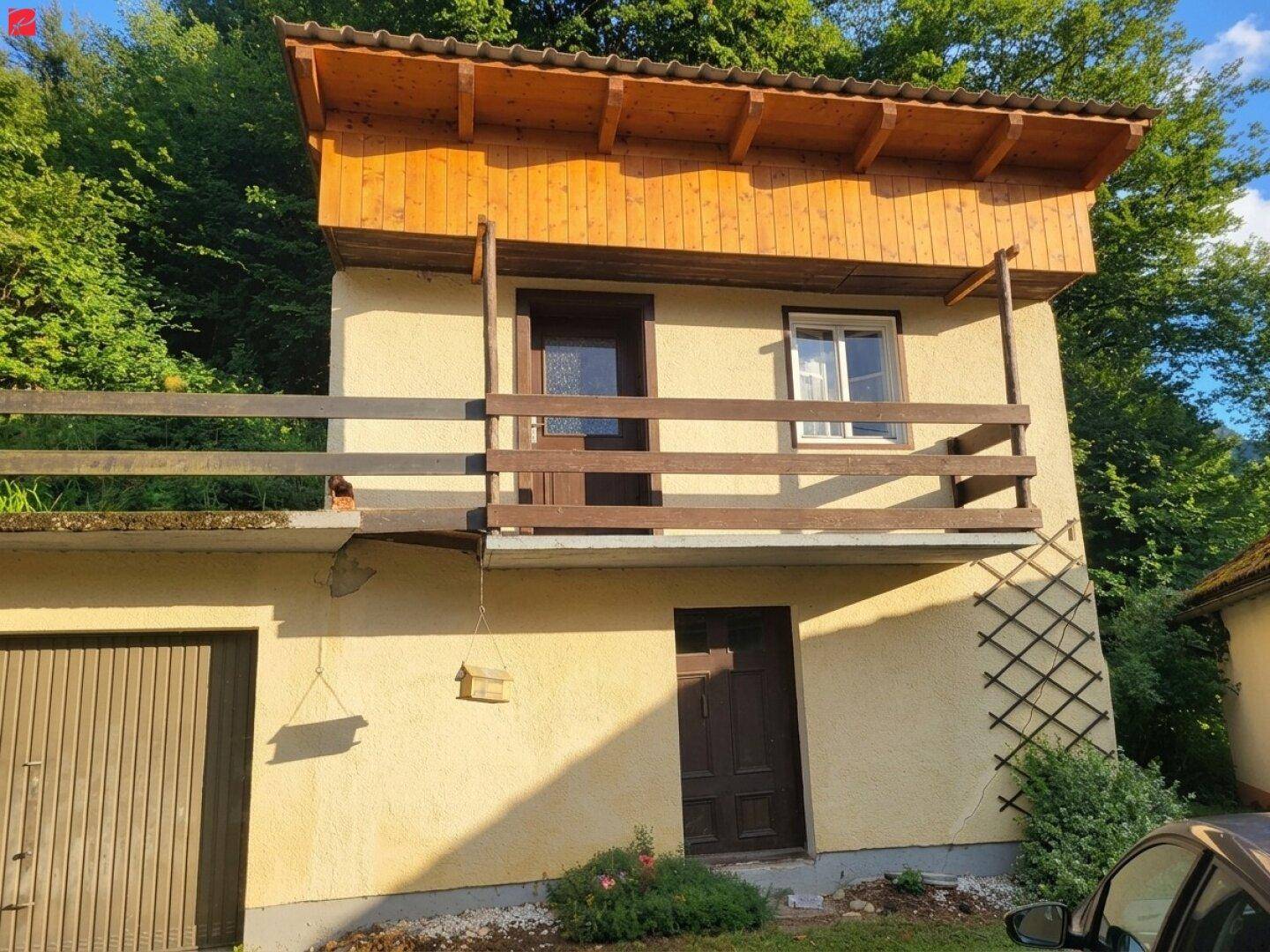 Fassade des Hauses mit Holzbalkon, Tür und Fenster, umgeben von üppigem Grün.