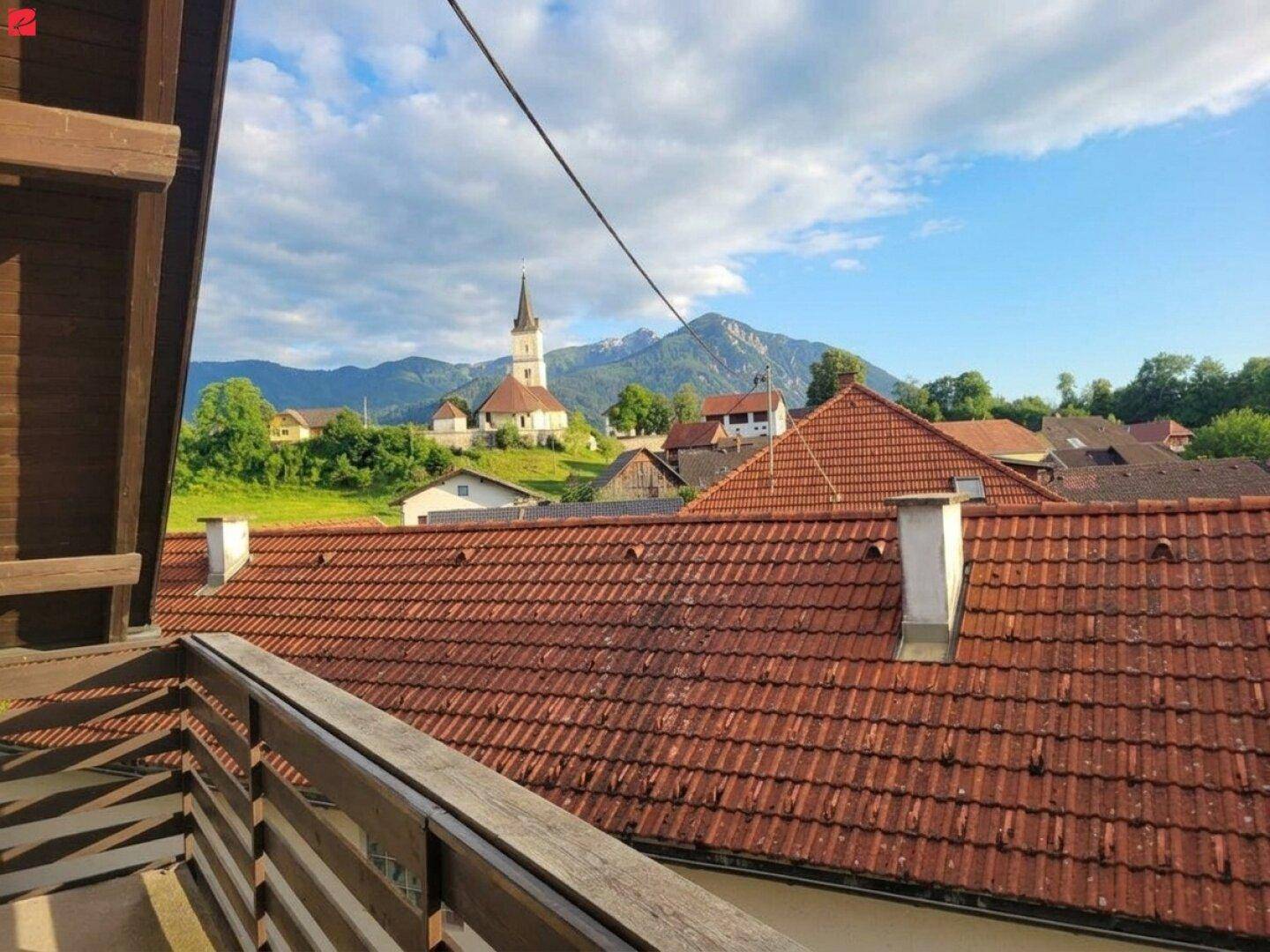 Panoramablick über ein Dorf mit Kirche und Bergen im Hintergrund unter blauem Himmel.