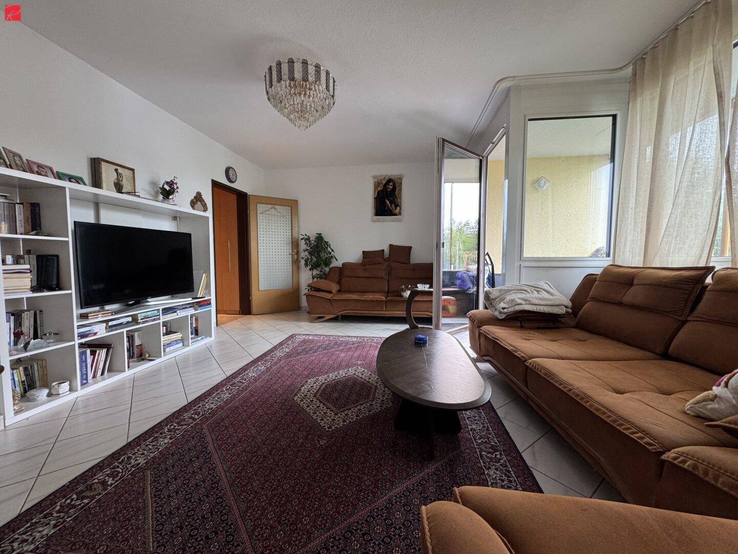 Helles Wohnzimmer mit braunen Sofas, Fliesenboden, großem Teppich und Zugang zu einem Balkon.