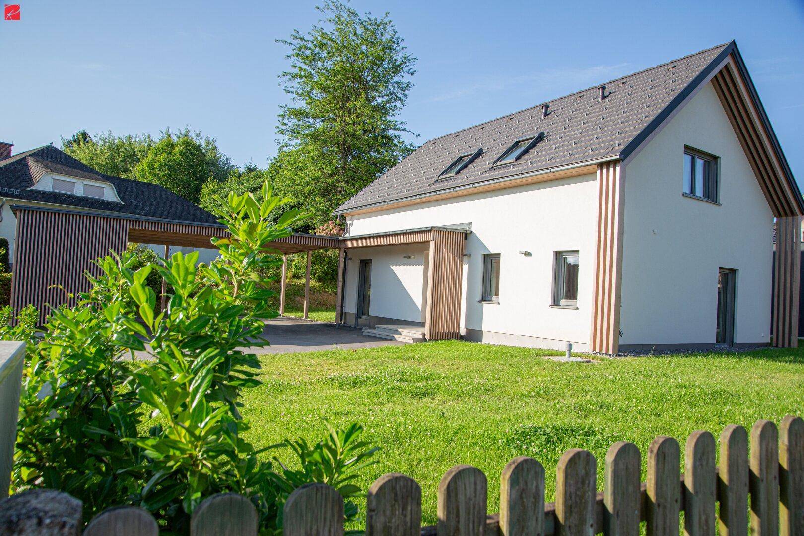 Modernes Einfamilienhaus mit weißer Fassade, Holzakzenten, Carport und einem gepflegten Rasenbereich.