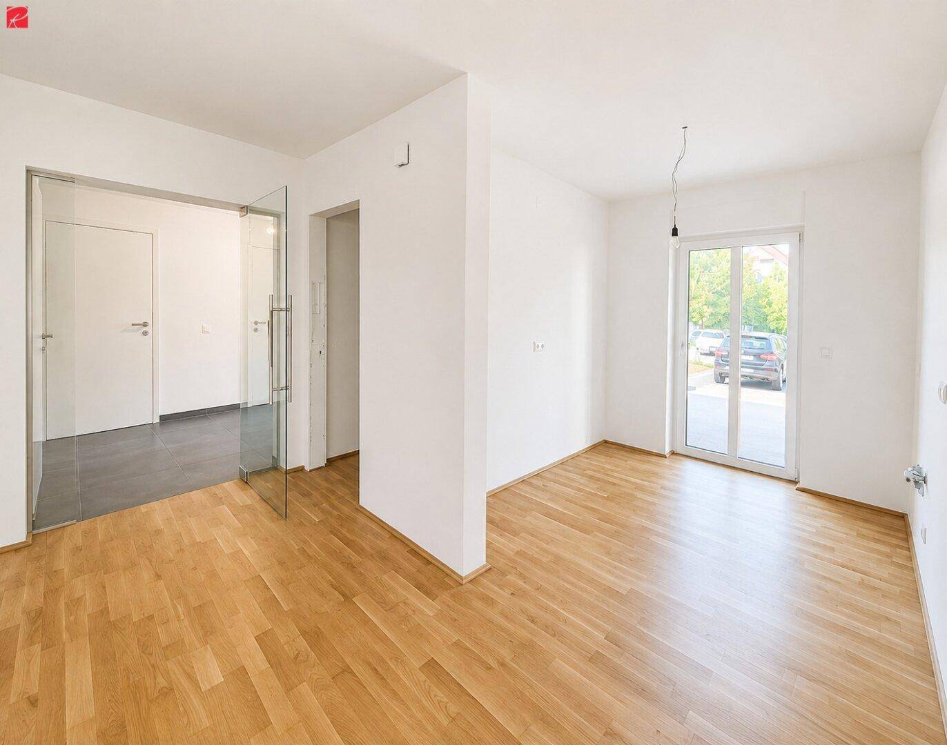 Geräumiger, heller Wohnraum mit Holzfußboden, weißen Wänden und Zugang zu einem Außenbereich.