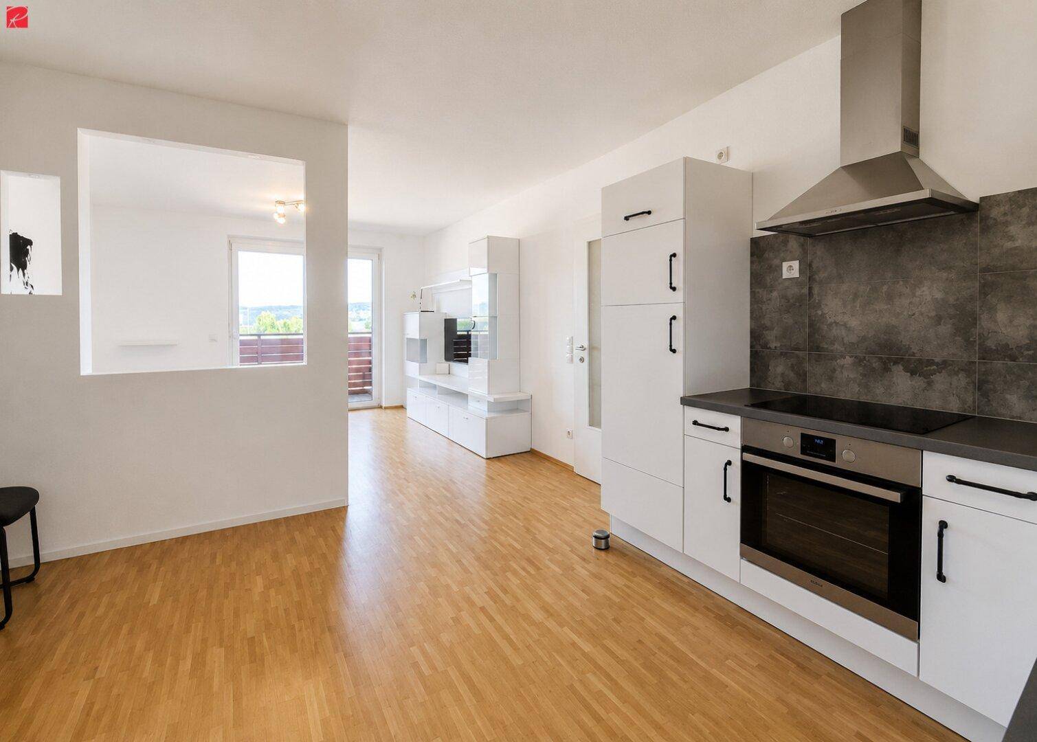 Offener Wohnbereich mit moderner Einbauküche, hellem Holzboden und Zugang zum Balkon.