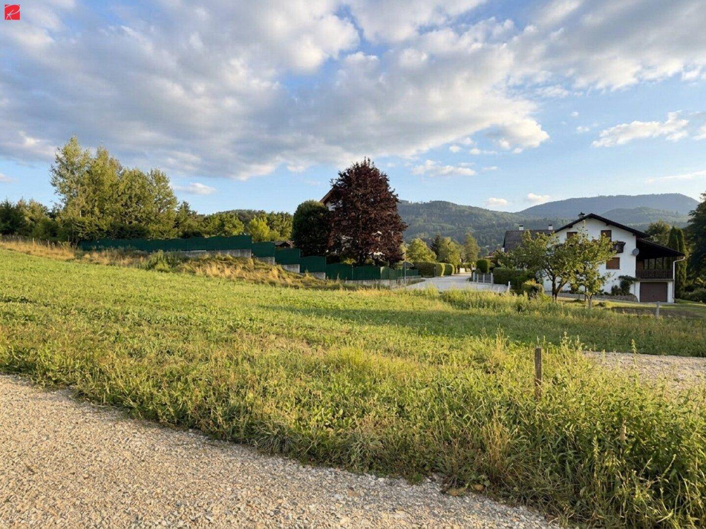Grünes Feld mit Häusern und Bäumen im Hintergrund, teilweise bewölkter Himmel.