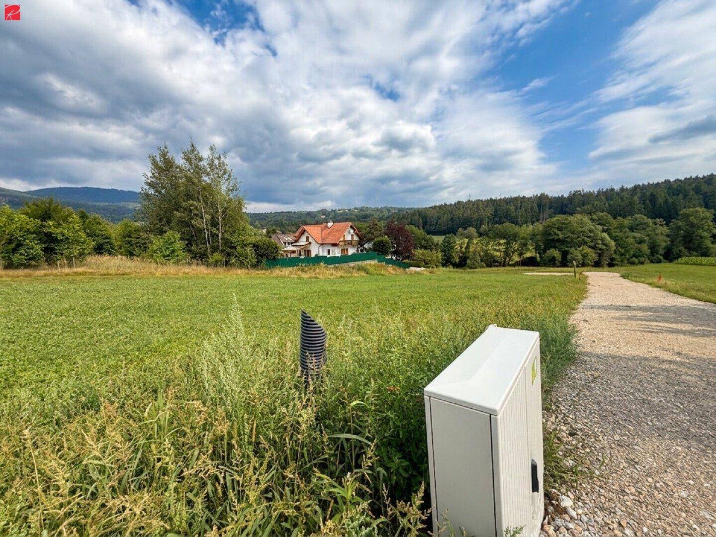 Grüne Wiese mit einem Feldweg und einem Versorgungsanschluss, im Hintergrund ein Haus und Wälder.