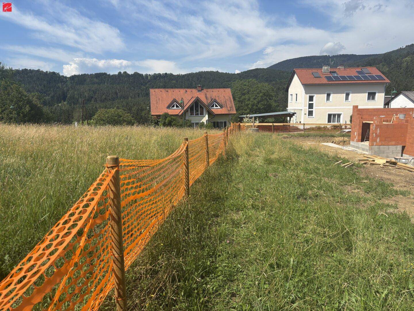 Blick auf ein Baugrundstück mit angrenzenden Häusern und bewaldeten Hügeln im Hintergrund an einem sonnigen Tag.