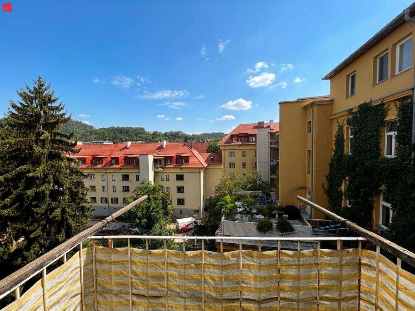 Blick vom Balkon auf umliegende Gebäude mit roten Dächern und grüne Bäume unter blauem Himmel.