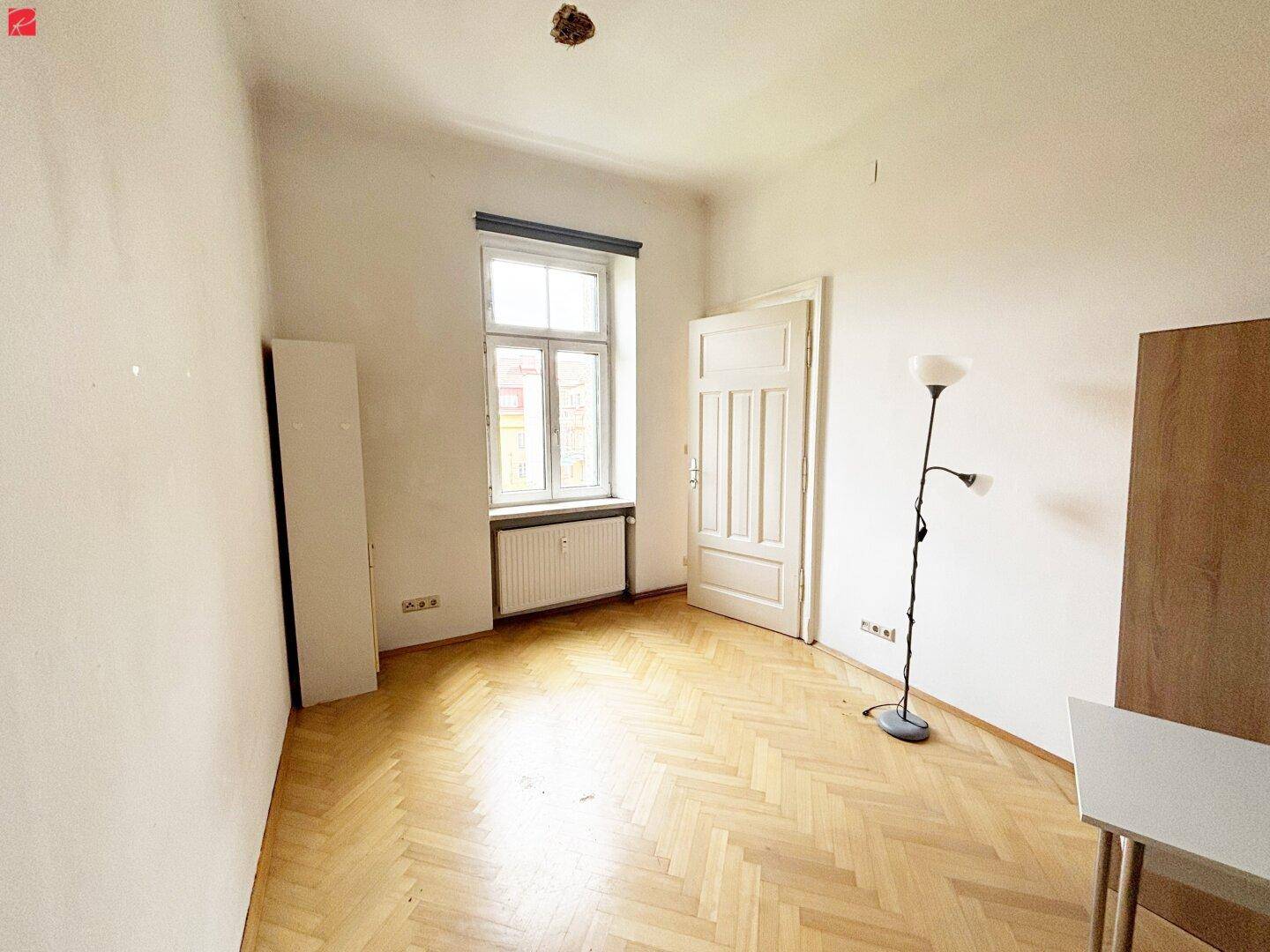 Leeres Zimmer mit Fischgrätparkett, großem Fenster und weißer Tür.
