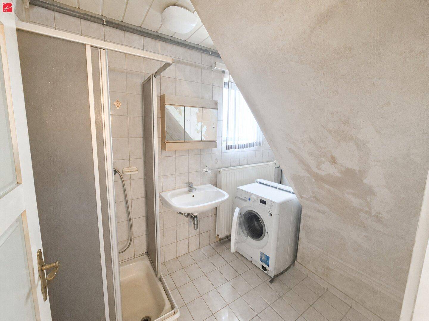 Kleines Badezimmer mit Dusche, Waschbecken und Waschmaschine unter der Dachschräge.