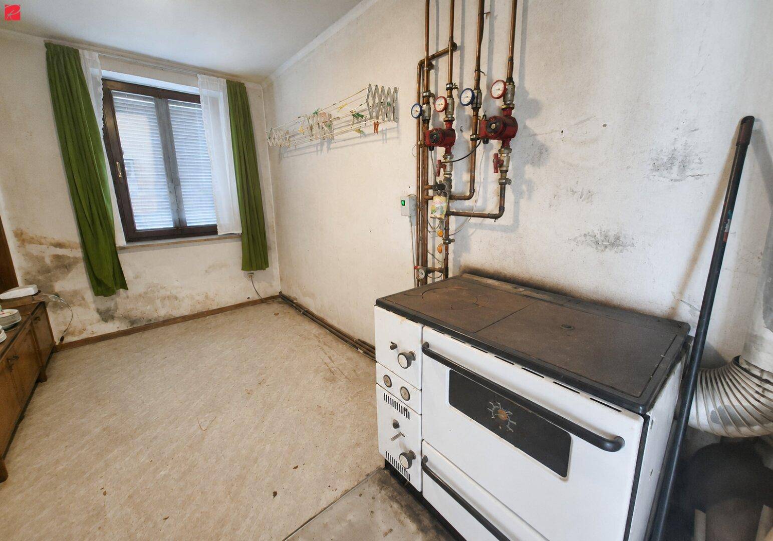 Funktionaler Raum mit altem Ofen, sichtbaren Rohren und Fenster, renovierungsbedürftig.