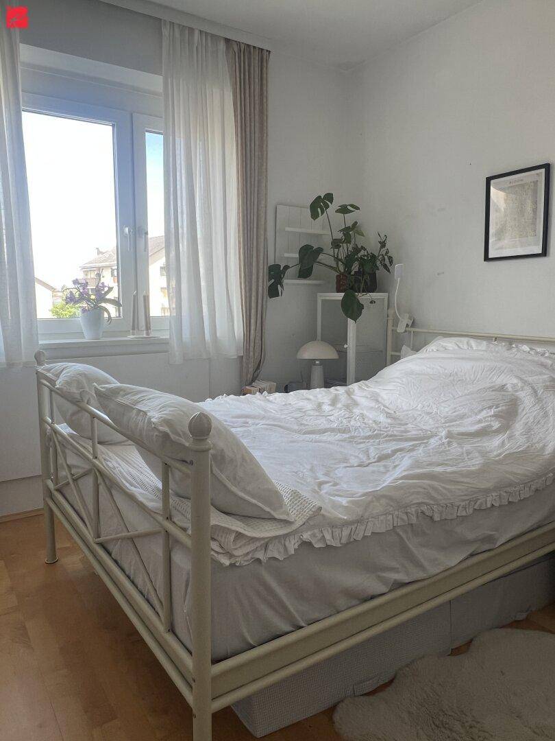 Helles Schlafzimmer mit weißem Metallbett, Holzboden und einem Fenster, das viel Tageslicht hereinlässt.