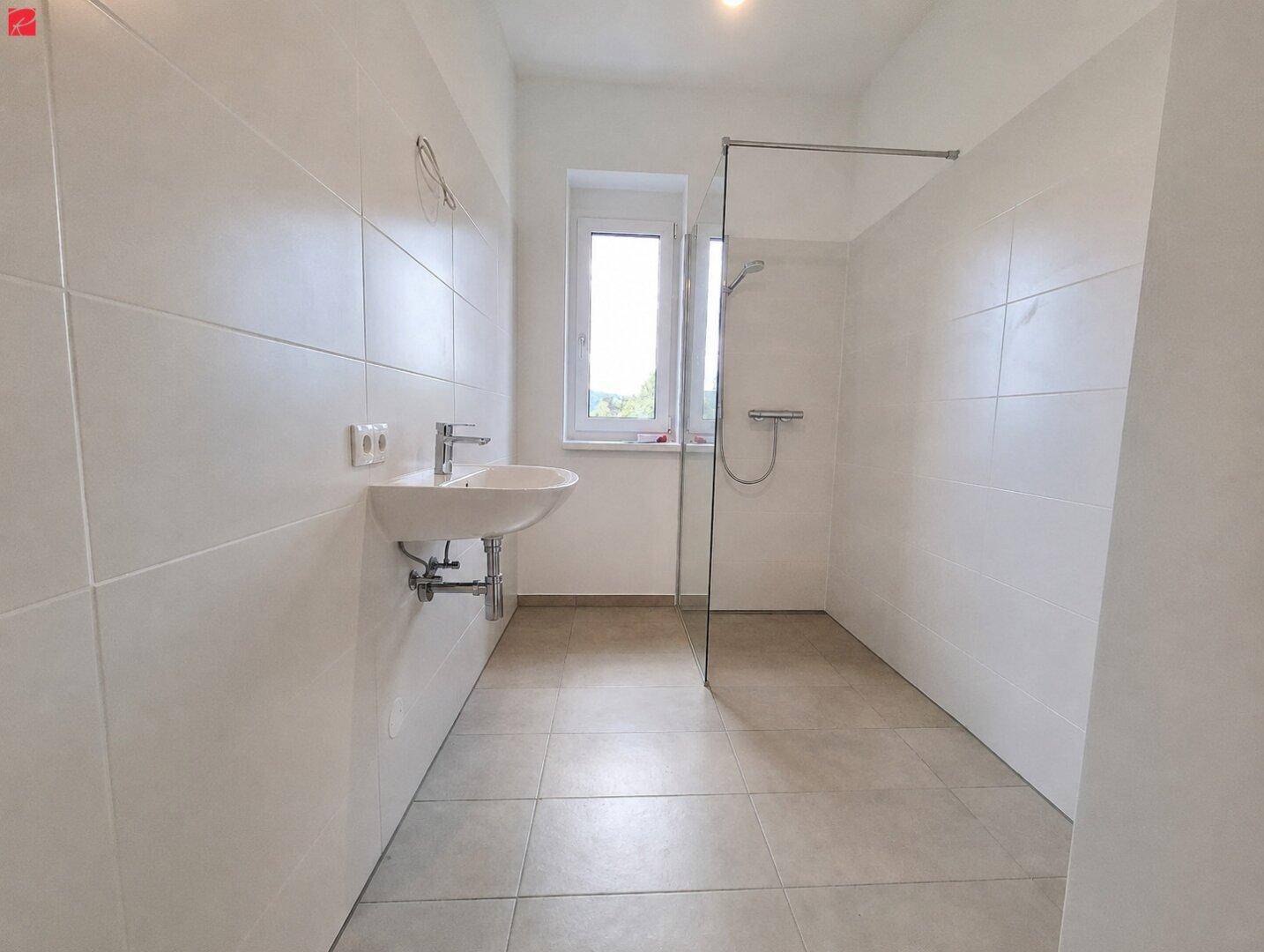 Modernes Badezimmer mit großformatigen weißen Fliesen, ebenerdiger Dusche und Fenster für Tageslicht.