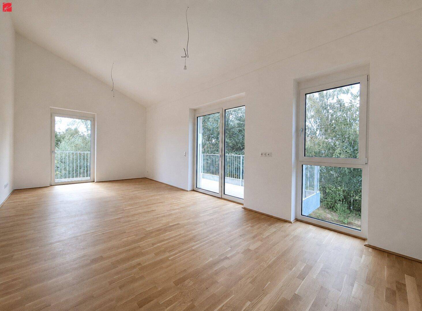Heller, leerer Wohnraum mit Holzparkettboden, großen Fenstern und Zugang zum Balkon.