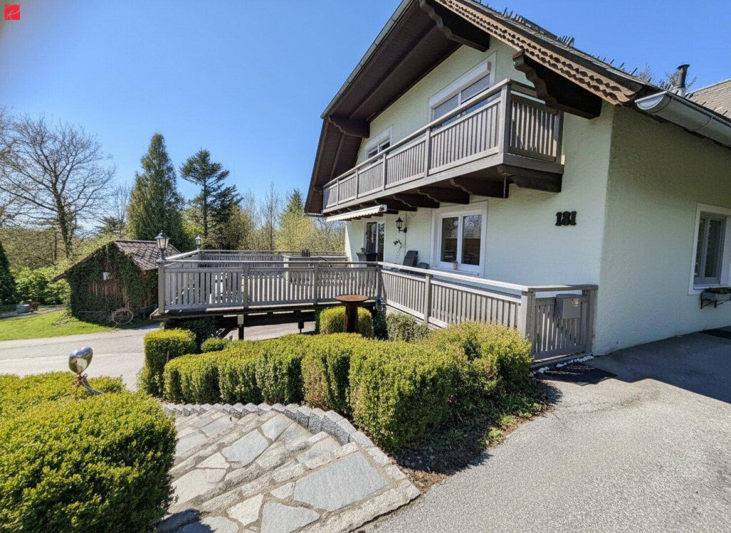 Charmantes Haus mit traditionellem Holzbalkon und gepflegter Gartenanlage unter blauem Himmel.
