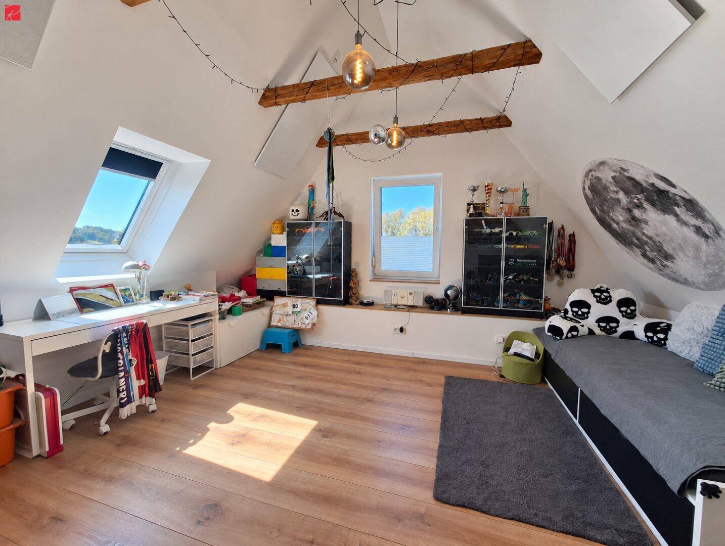 Helles Schlafzimmer im Dachgeschoss mit Holzbalken, Schreibtisch und großem Fenster mit Ausblick.