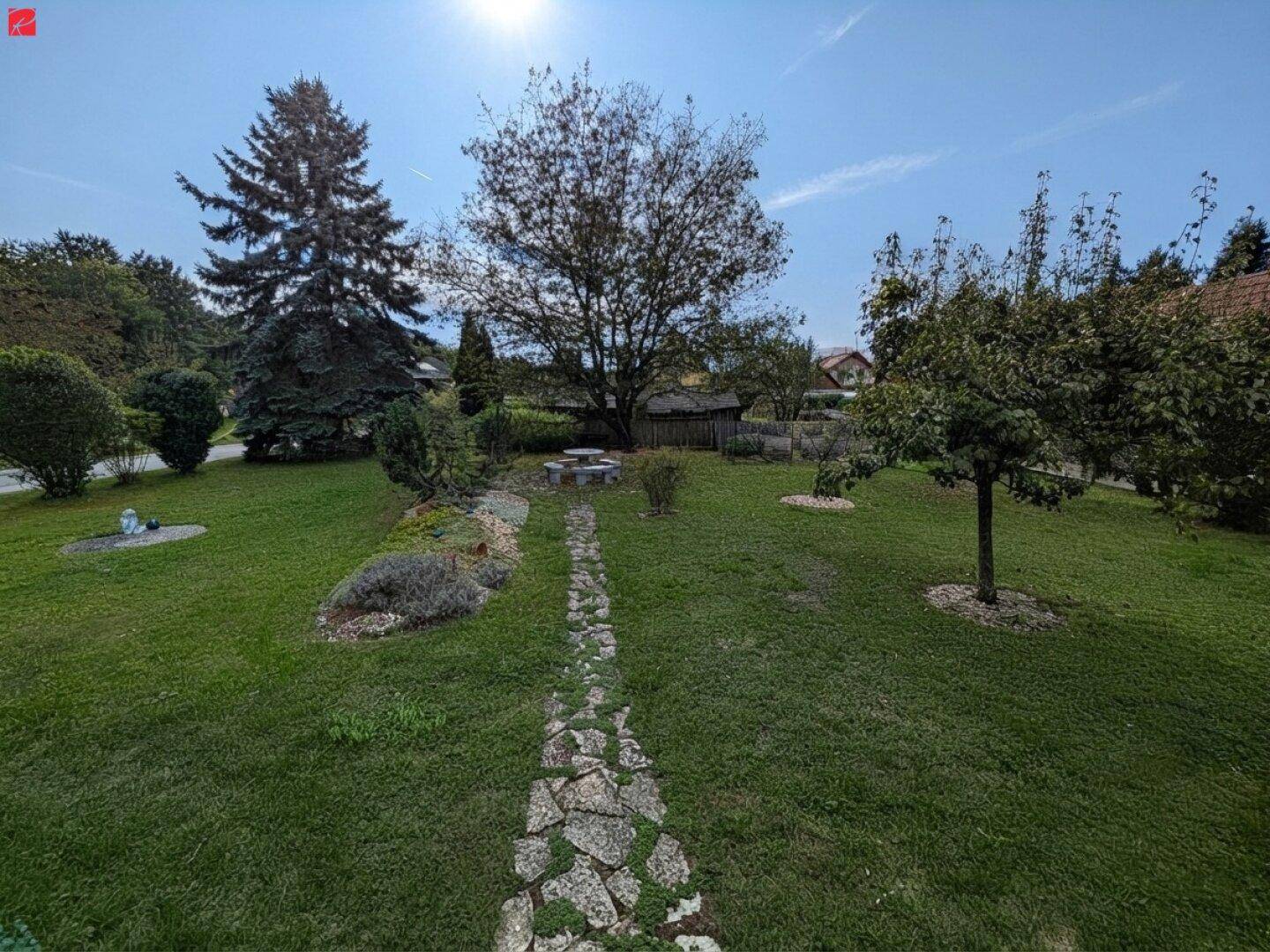 Weitläufiger Garten mit altem Baumbestand, Rasenflächen und einem Steinweg unter blauem Himmel.