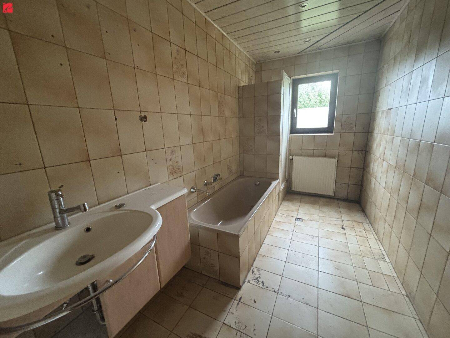 Badezimmer mit beigefarbenen Fliesen, Badewanne und Waschbecken, das eine Modernisierung benötigt.