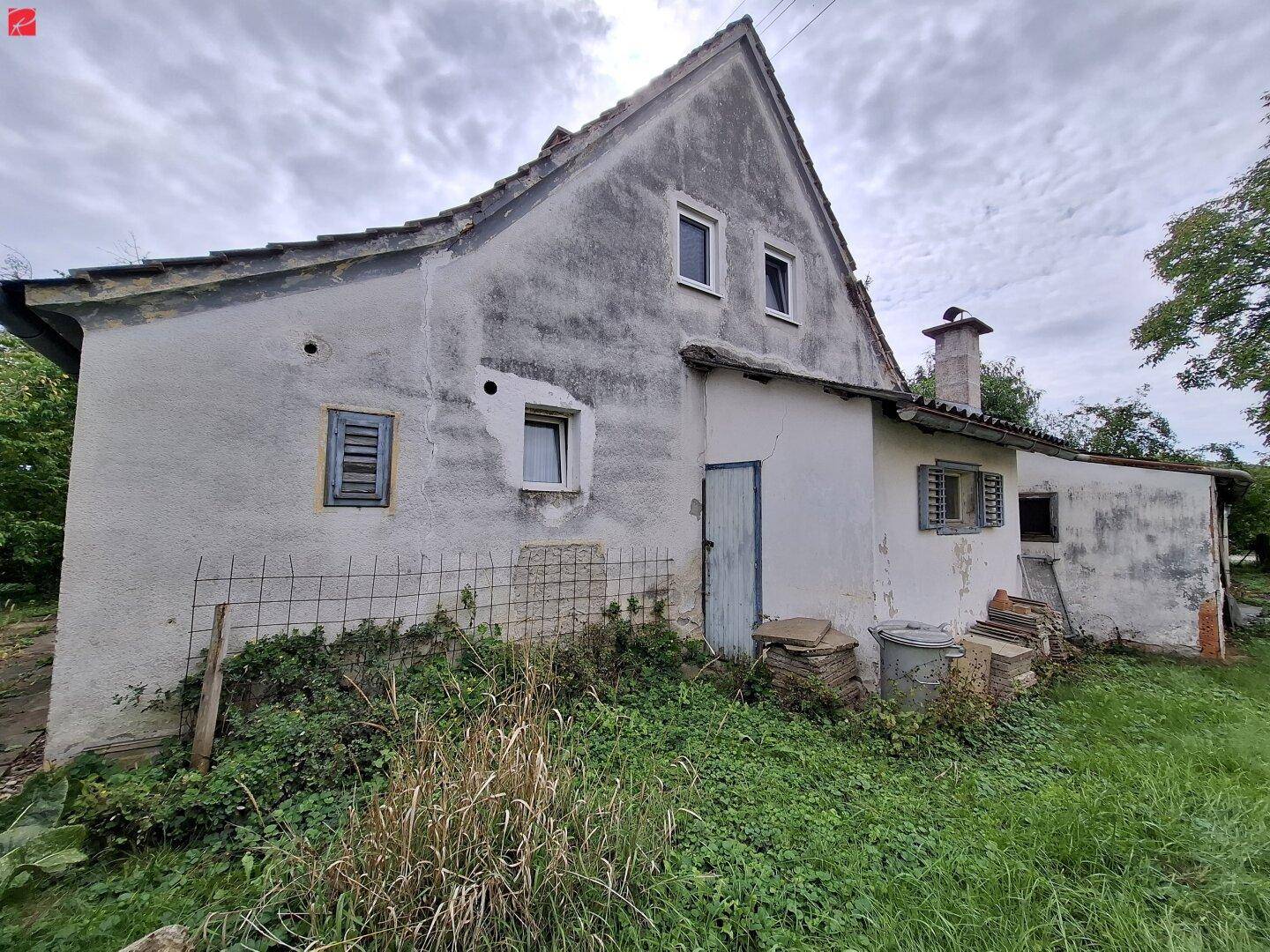 Rückansicht eines älteren Hauses mit verwitterter Fassade und einem kleinen Anbau im Grünen.