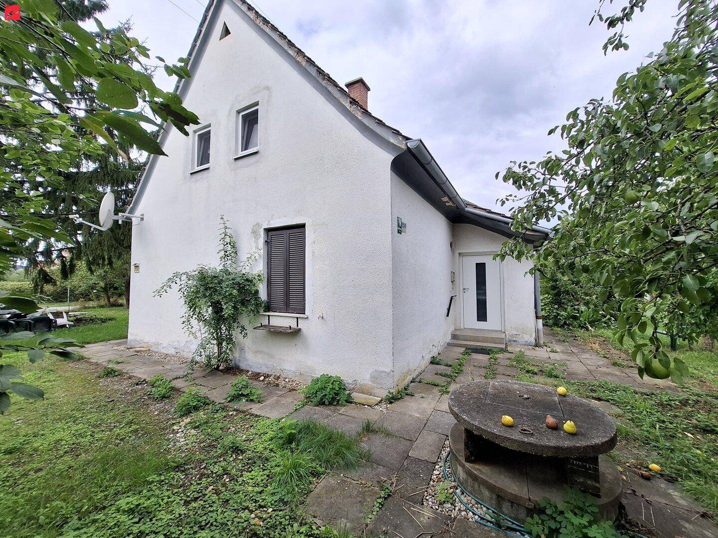 Vorderansicht eines einfachen Hauses mit Garten, Obstbäumen und einem steinernen Gartentisch.