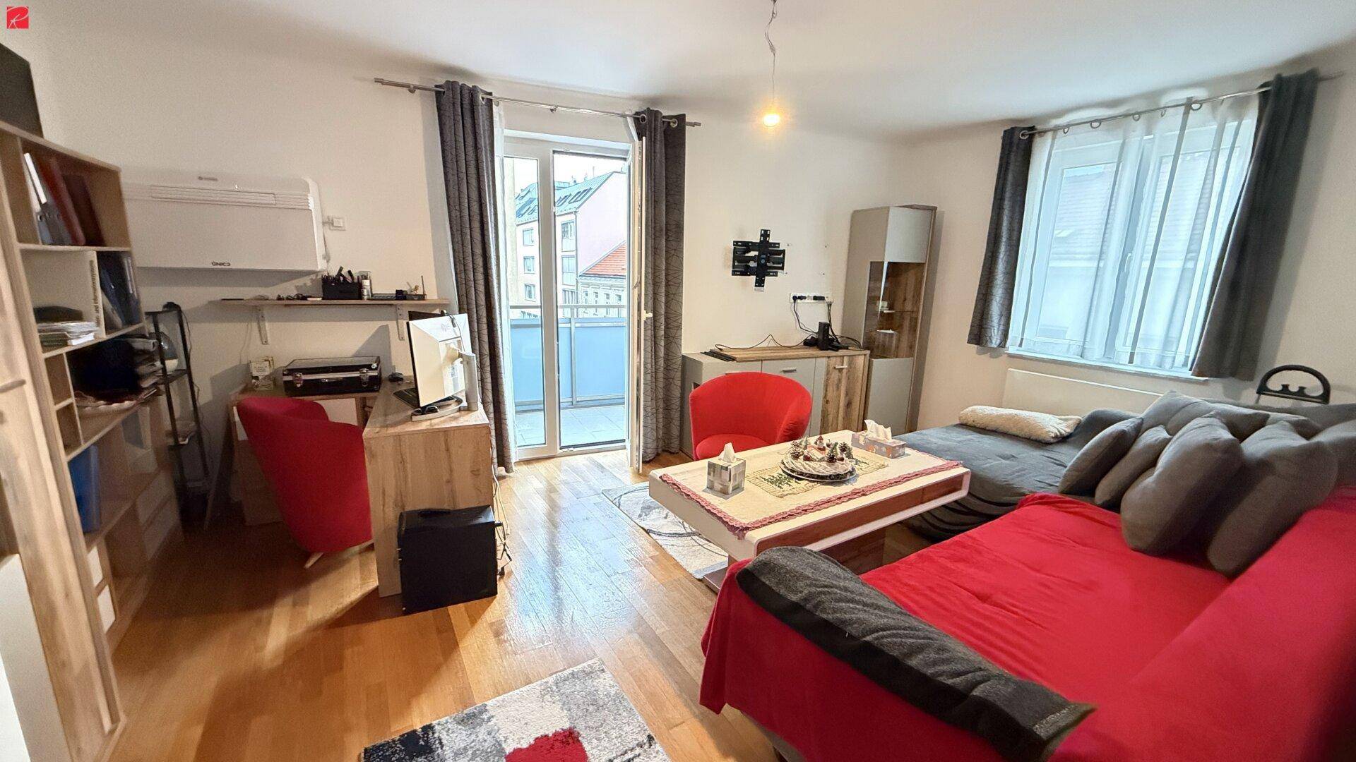Helles Wohnzimmer mit Holzboden, rotem Sofa, Schreibtisch und Zugang zum Balkon.