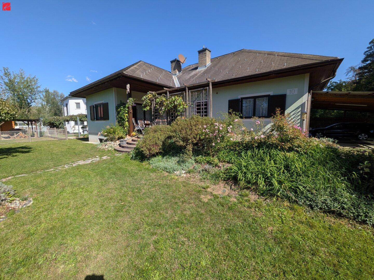 Weitläufiger Gartenbereich vor dem Haus mit Carport und üppiger Bepflanzung unter blauem Himmel.