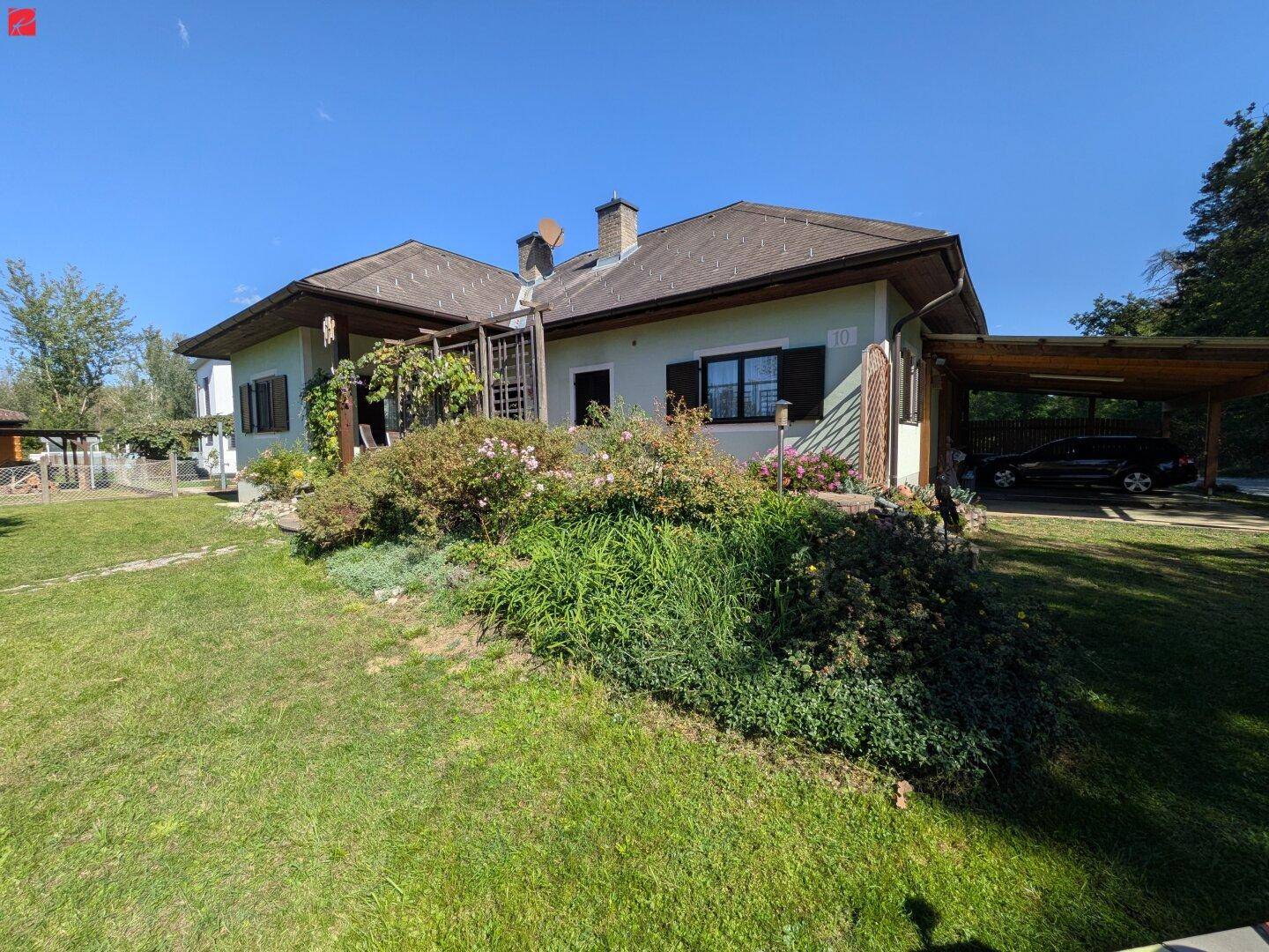 Hausansicht mit großem Garten, überdachter Terrasse und einem Carport für Fahrzeuge.