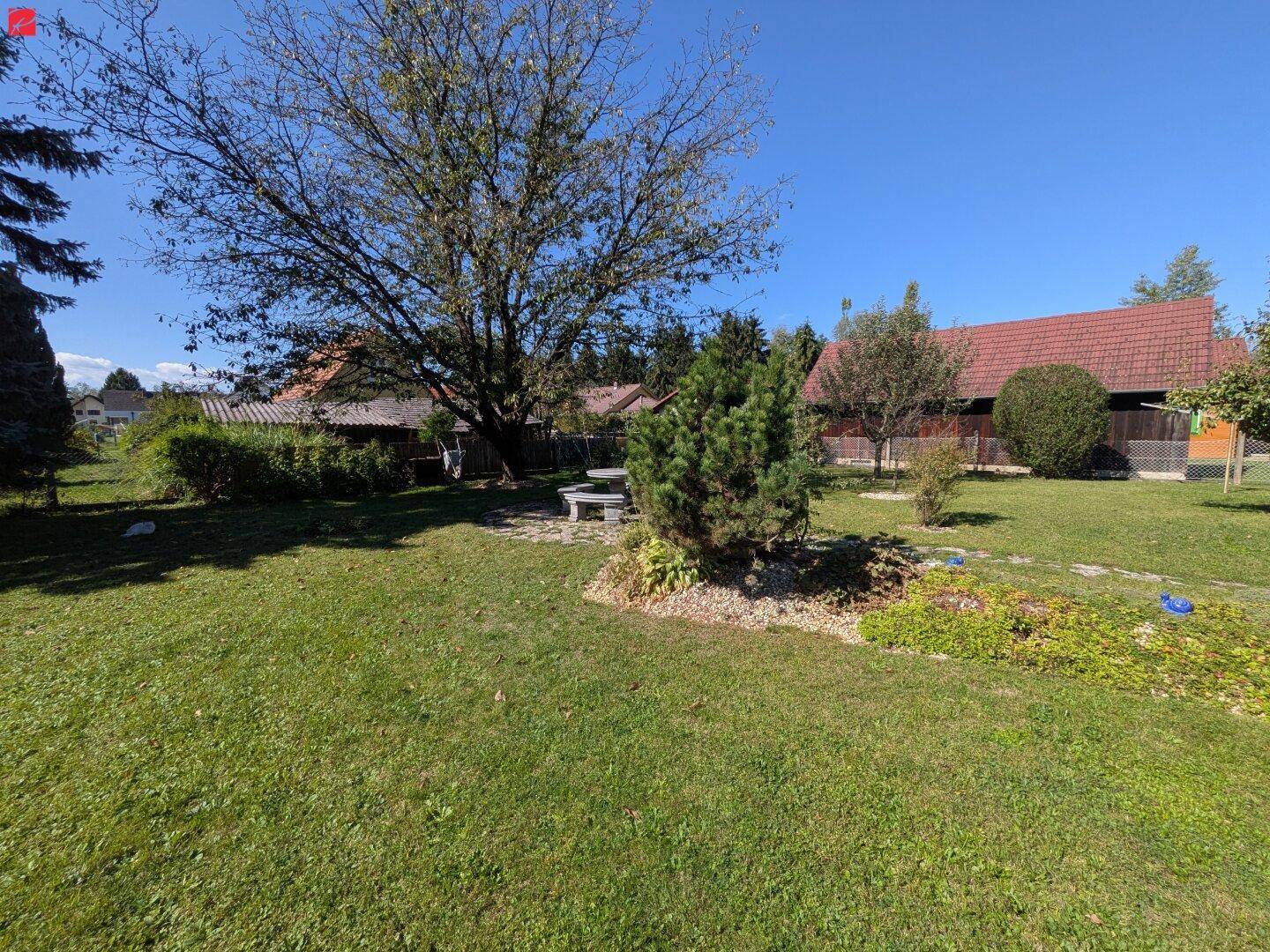 Gepflegter Garten mit Rasen, Bäumen, Sträuchern und einem steinernen Sitzbereich im Hintergrund.