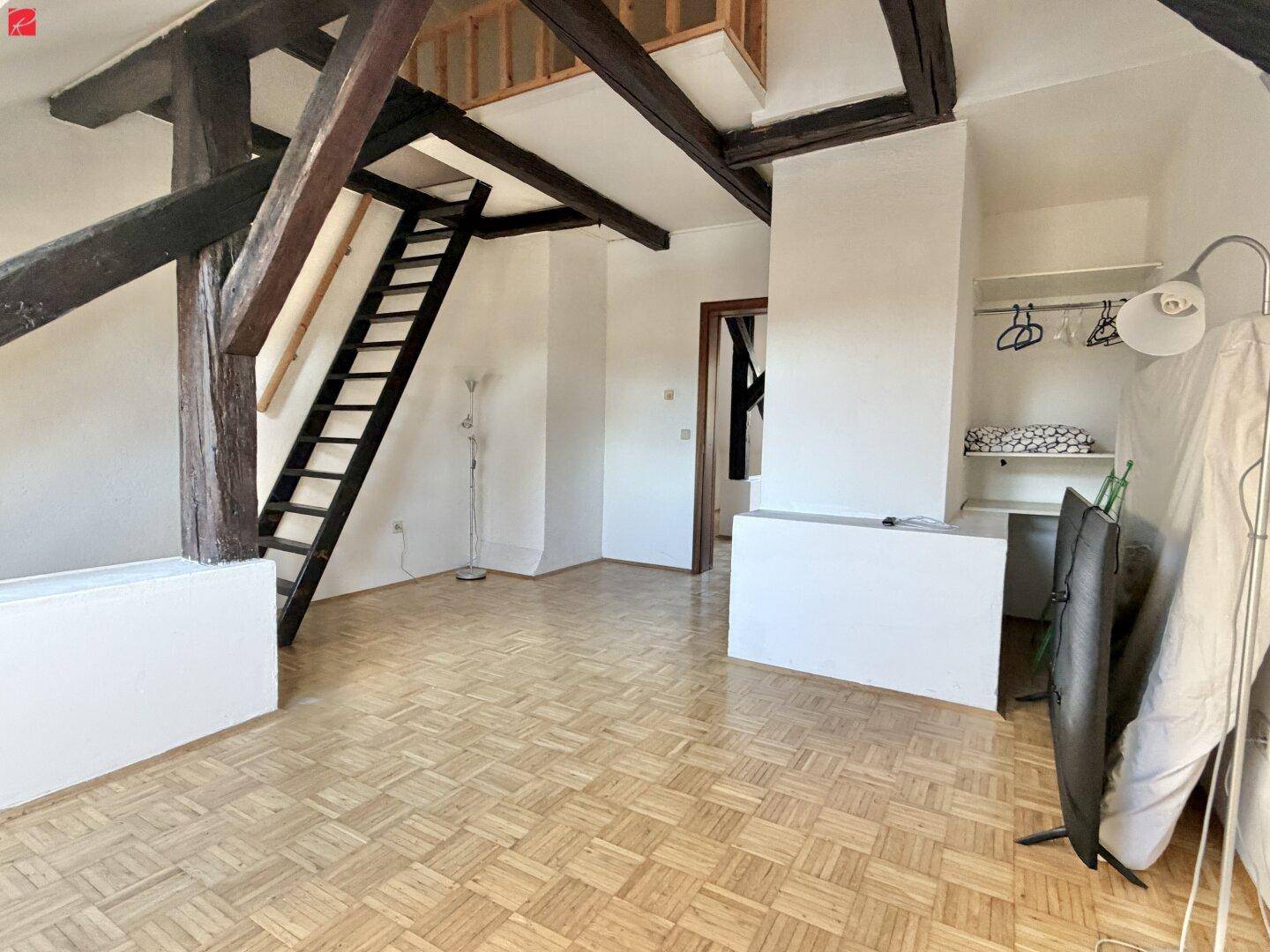 Geräumiges Dachgeschosszimmer mit sichtbaren Holzbalken und offener Treppe zu einer weiteren Ebene.