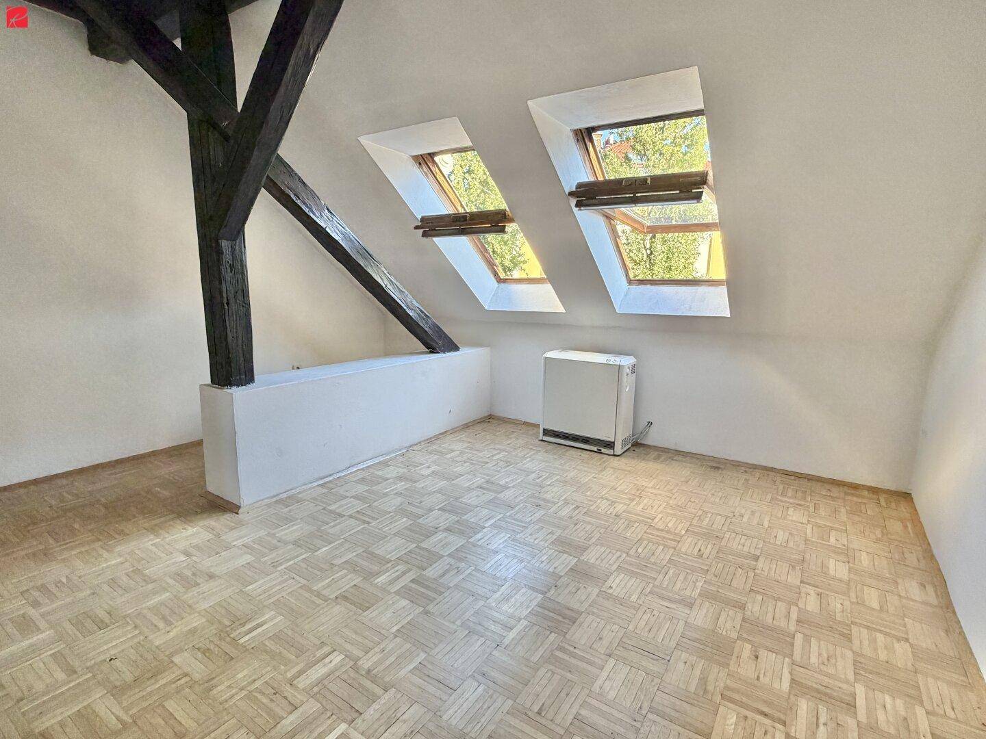 Lichtdurchfluteter Dachbodenraum mit sichtbaren Holzbalken, Parkettboden und zwei Dachfenstern.