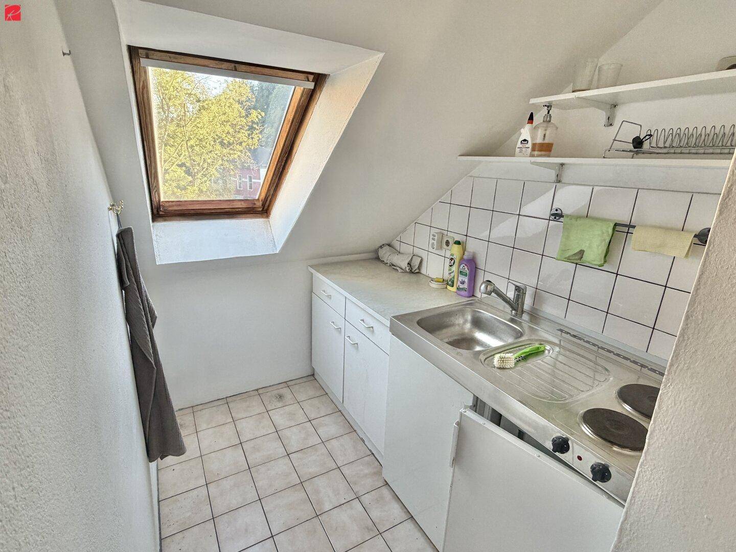 Kleine Küchenzeile im Dachgeschoss mit Spüle, Kochfeld, weißen Schränken und Dachfenster.