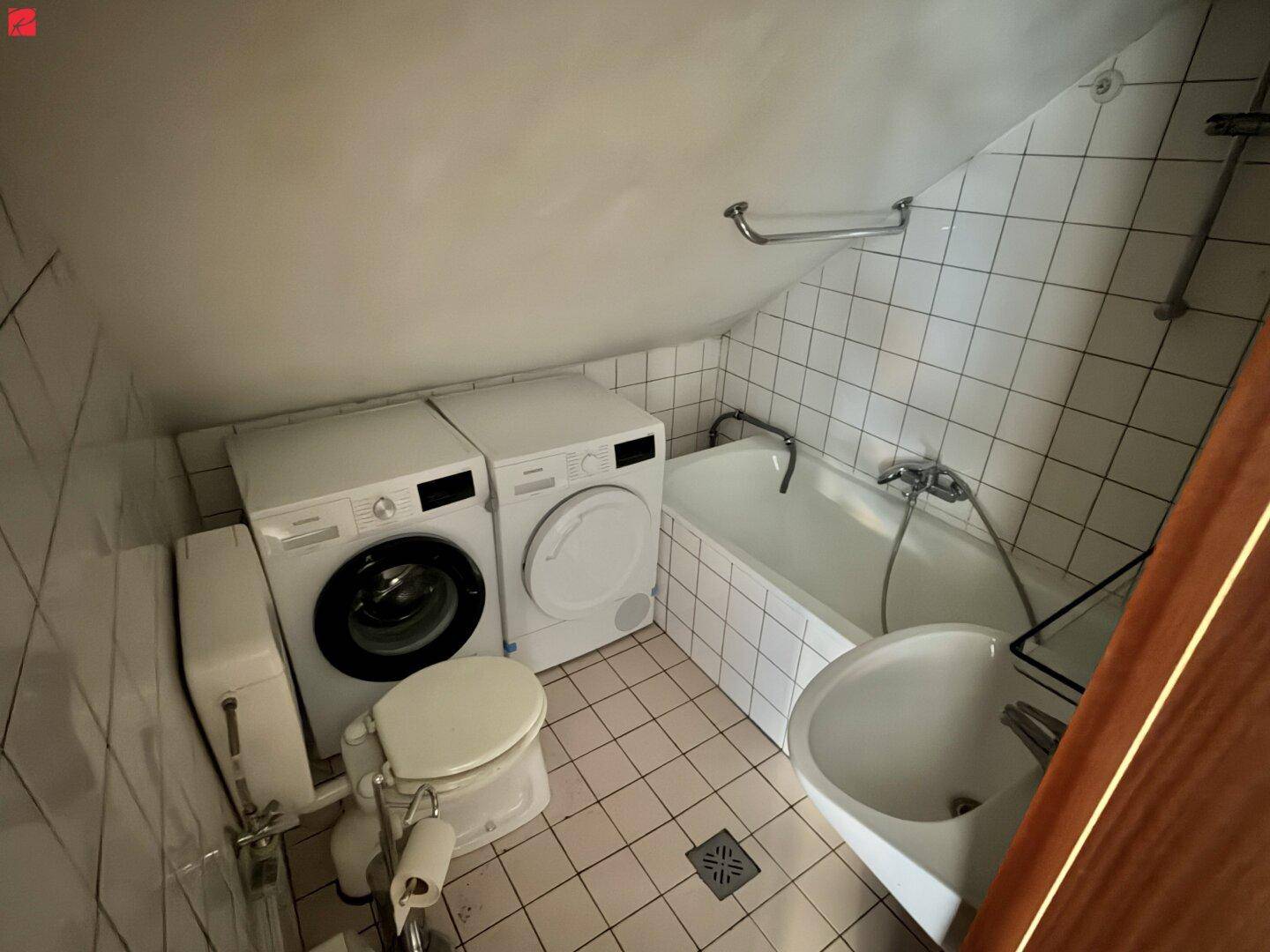Kleines Badezimmer mit Badewanne, WC, Waschmaschine, Trockner und Dachschräge.