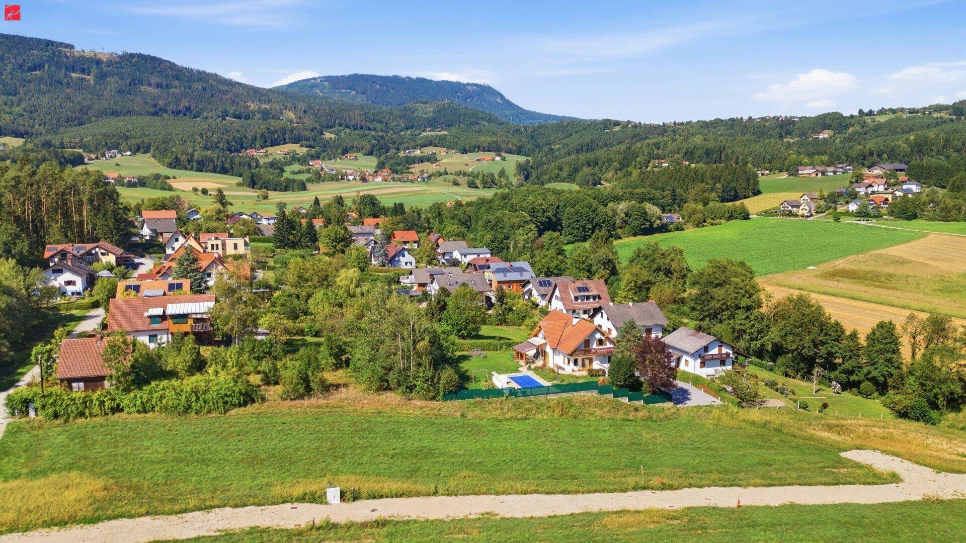 Panoramablick auf eine Wohnsiedlung in einer grünen, hügeligen Landschaft mit Bergen im Hintergrund.